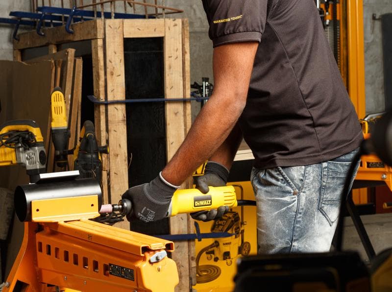 Persona usando amoladora DEWALT DWE4887N en metal, taller con otras herramientas amarillas visibles.