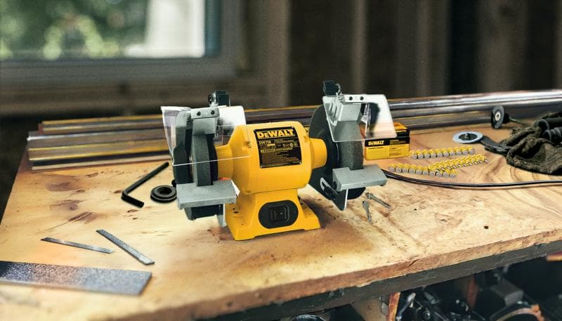 Un esmeril de banco DEWALT, modelo DW758, está colocado sobre una mesa de trabajo de madera. Alrededor hay varias herramientas y piezas metálicas, sugiriendo un espacio de trabajo activo. El esmeril es amarillo y tiene dos ruedas abrasivas con protectores de seguridad.