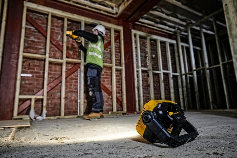 Luz de trabajo DEWALT DCL077 en el suelo, trabajador al fondo junto a estructura de madera.