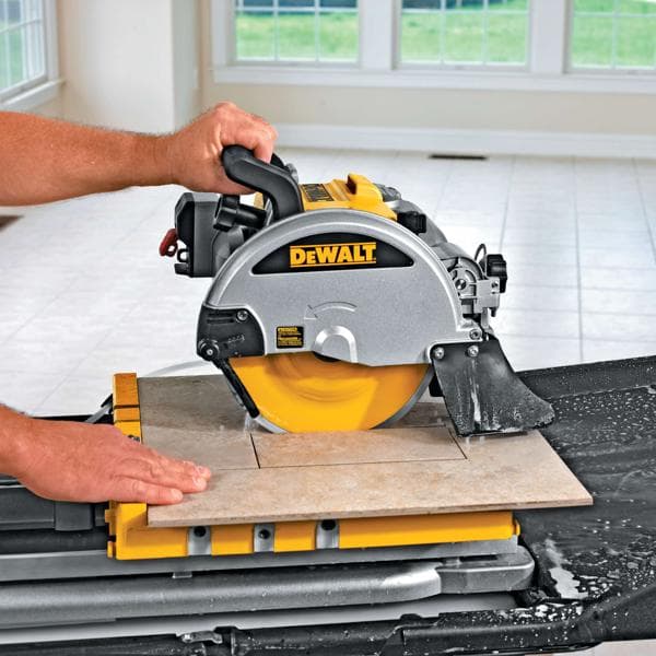 Vista de primer plano de una sierra para azulejos DEWALT en uso, cortando una baldosa de cerámica sobre una mesa de trabajo amarilla. Se observa agua alrededor de la zona de corte y las manos del operador guían la baldosa. La sierra se utiliza en una sala luminosa con grandes ventanas y suelo de baldosas.