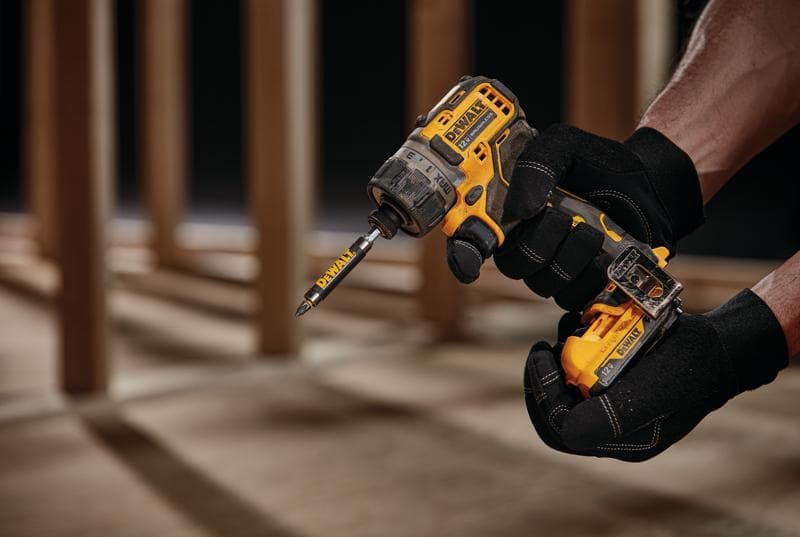 Vista en primer plano de una persona con guantes negros sosteniendo un taladro destornillador inalámbrico amarillo DEWALT con una broca acoplada, en un entorno de construcción con vigas de madera al fondo.