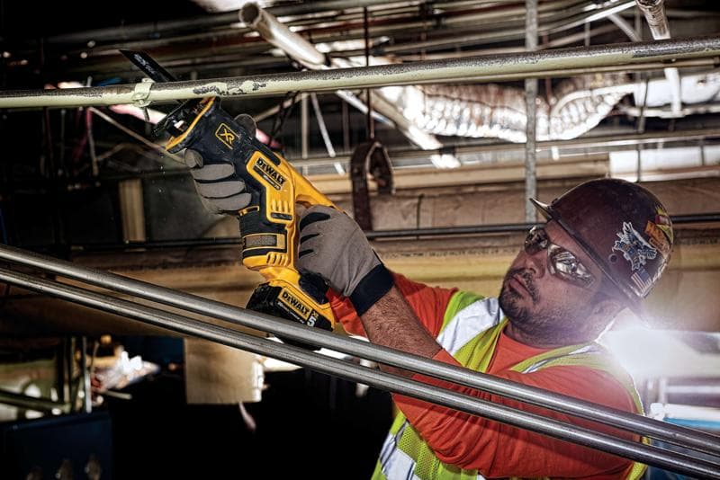 Una persona con guantes protectores, un chaleco de seguridad y un casco está usando una sierra recíproca inalámbrica DEWALT DCS367B para cortar una tubería de metal en un entorno industrial. El rostro de la persona está difuminado por privacidad.