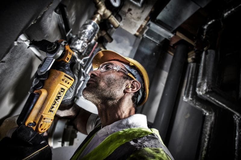 Trabajador corta tubo con DEWALT DWE315, lleva casco y chaleco en entorno industrial.