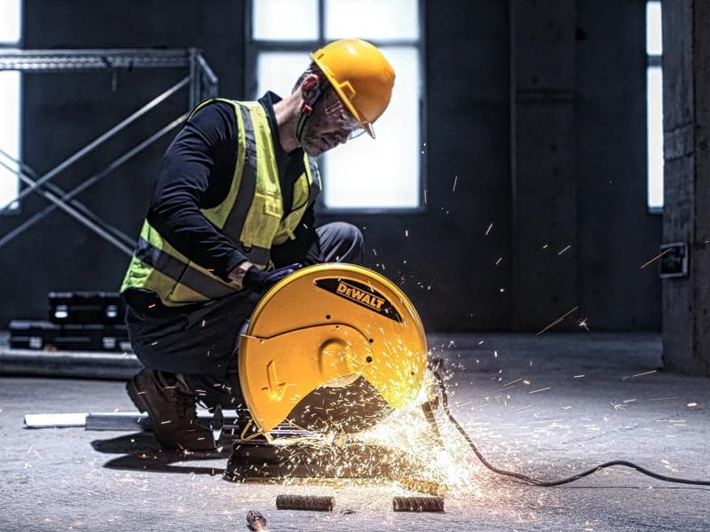 Una persona con casco de seguridad amarillo y chaleco reflectante utiliza una herramienta de corte DEWALT D28730 para cortar tubos de metal en un entorno industrial, con chispas visibles saliendo de la máquina.